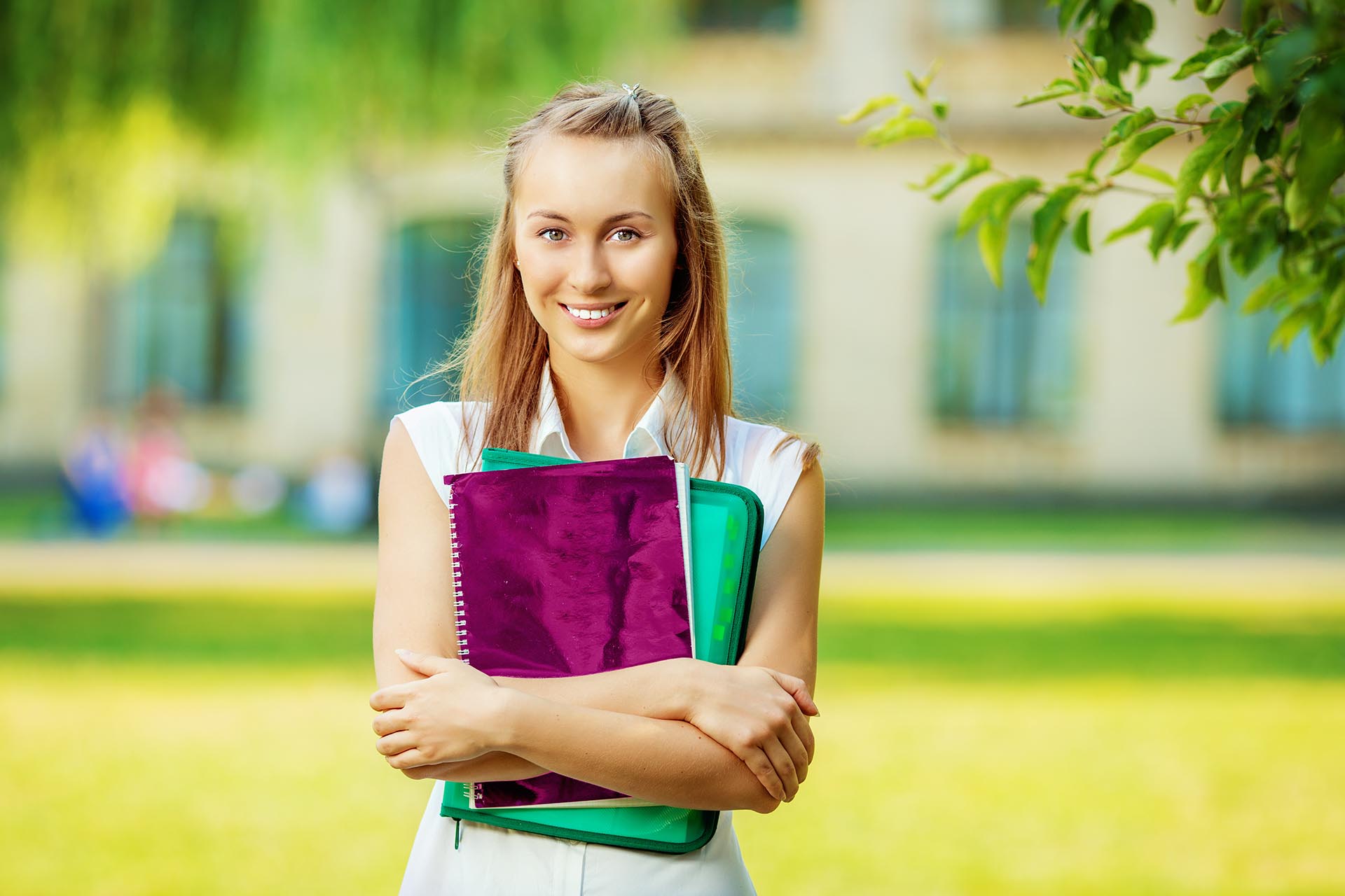 Student woman is standing with the folder and copy-book on the university campus background. Happy smart young woman is going to study outdoors in the park.